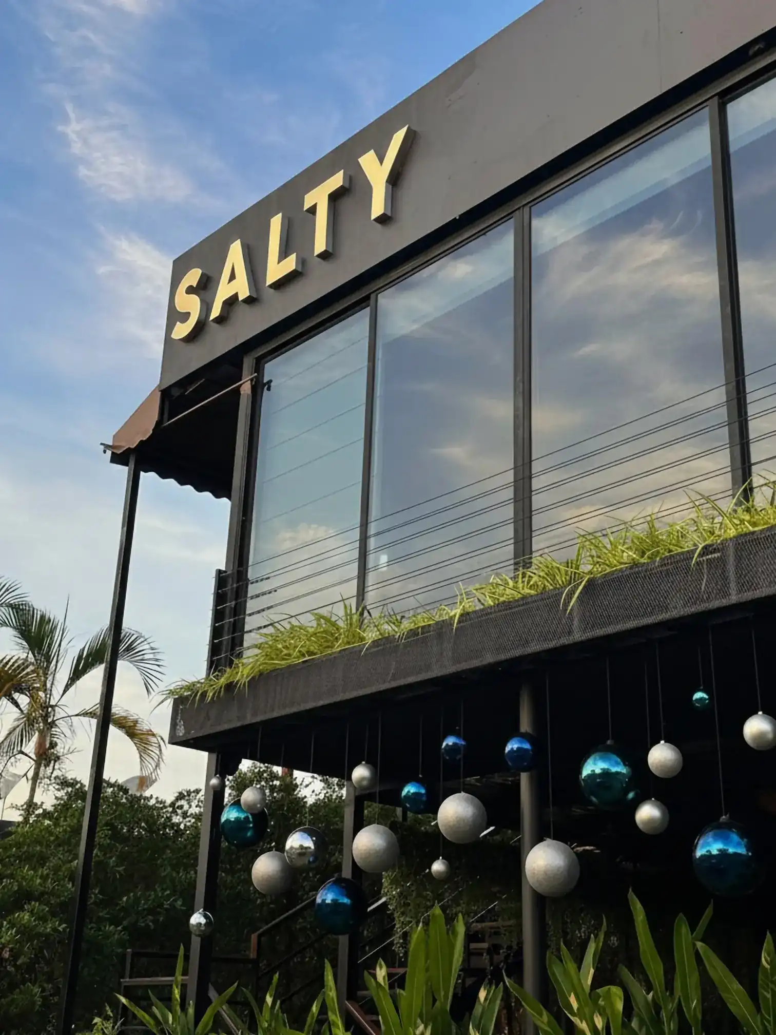 The brass SALTY sign above the two-storey glass front of the restaurant, with palms and blue and silver baubles under the terrace