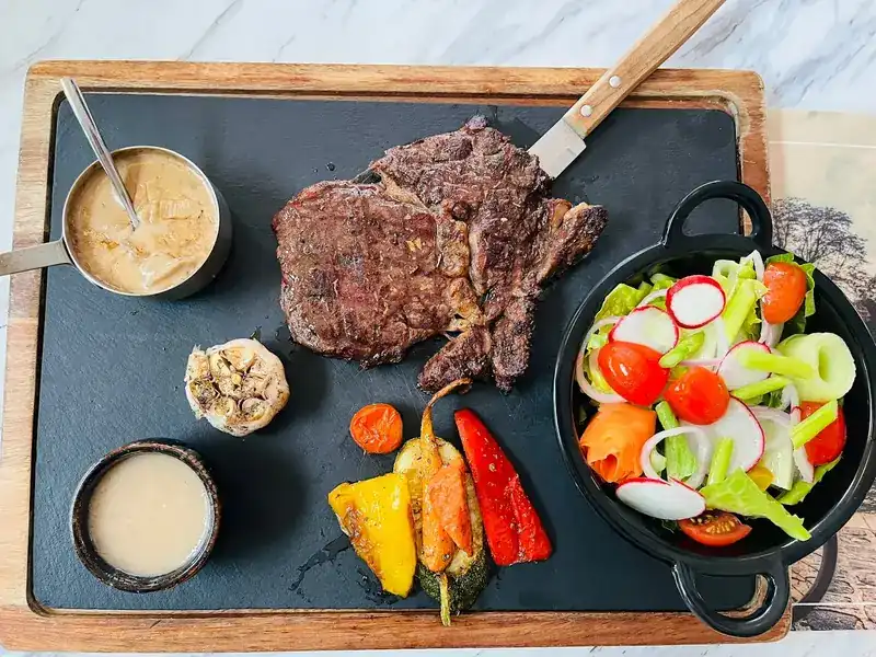 Steak on a slate board with grilled vegetables, salad, roasted garlic and two sauces
