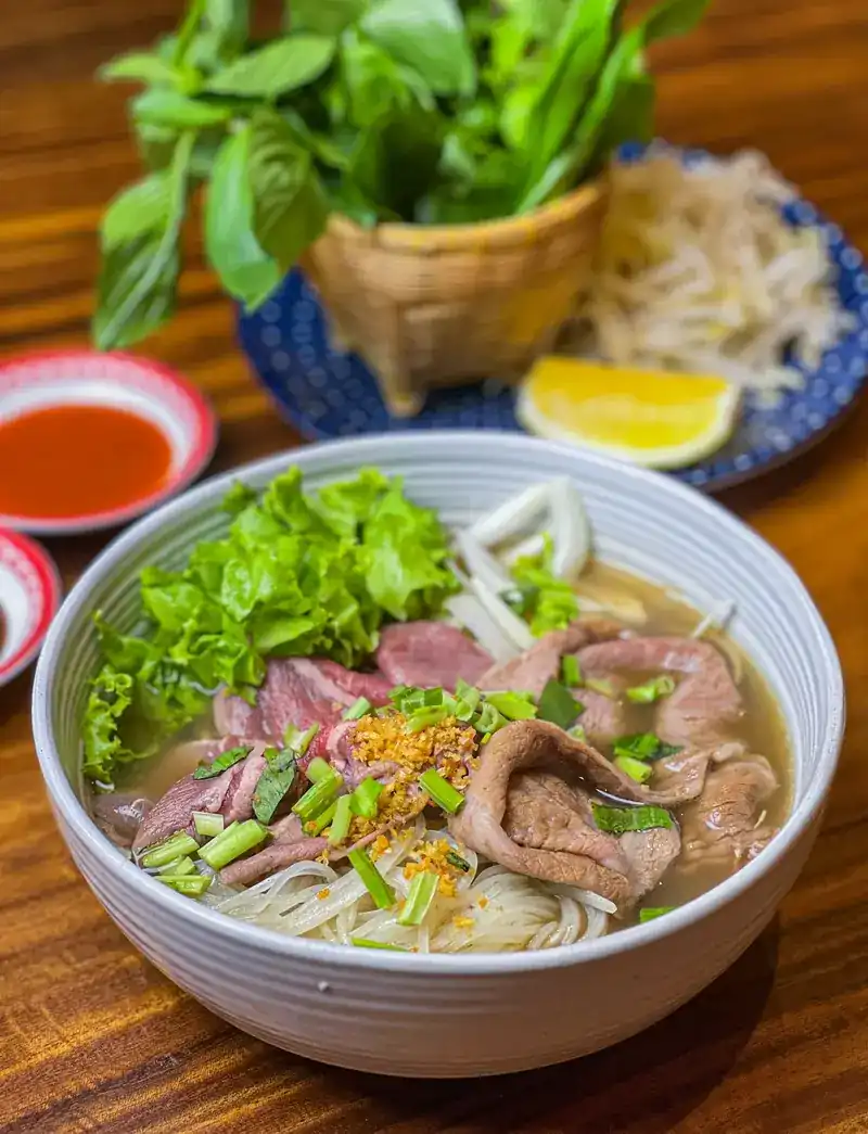 Pho with rare beef slices, rice noodles and fried garlic, with a basket of basil, bean sprouts, lime and chilli sauce on the side