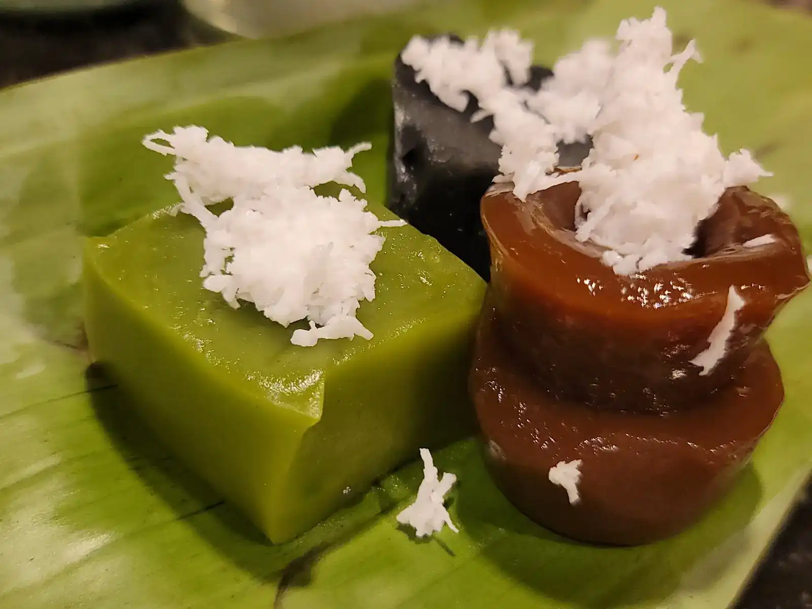 Pandan-green, black and dark-brown khanom topped with grated coconut, on banana leaf