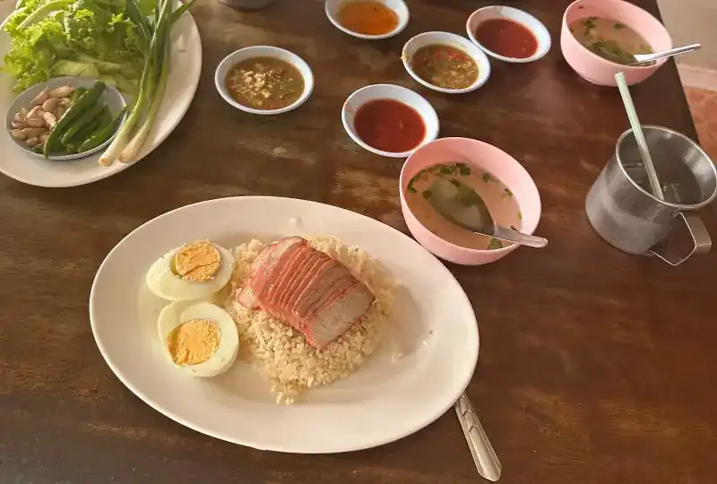 Red pork on rice with a halved boiled egg, pink bowls of soup and dipping sauces on a wooden table