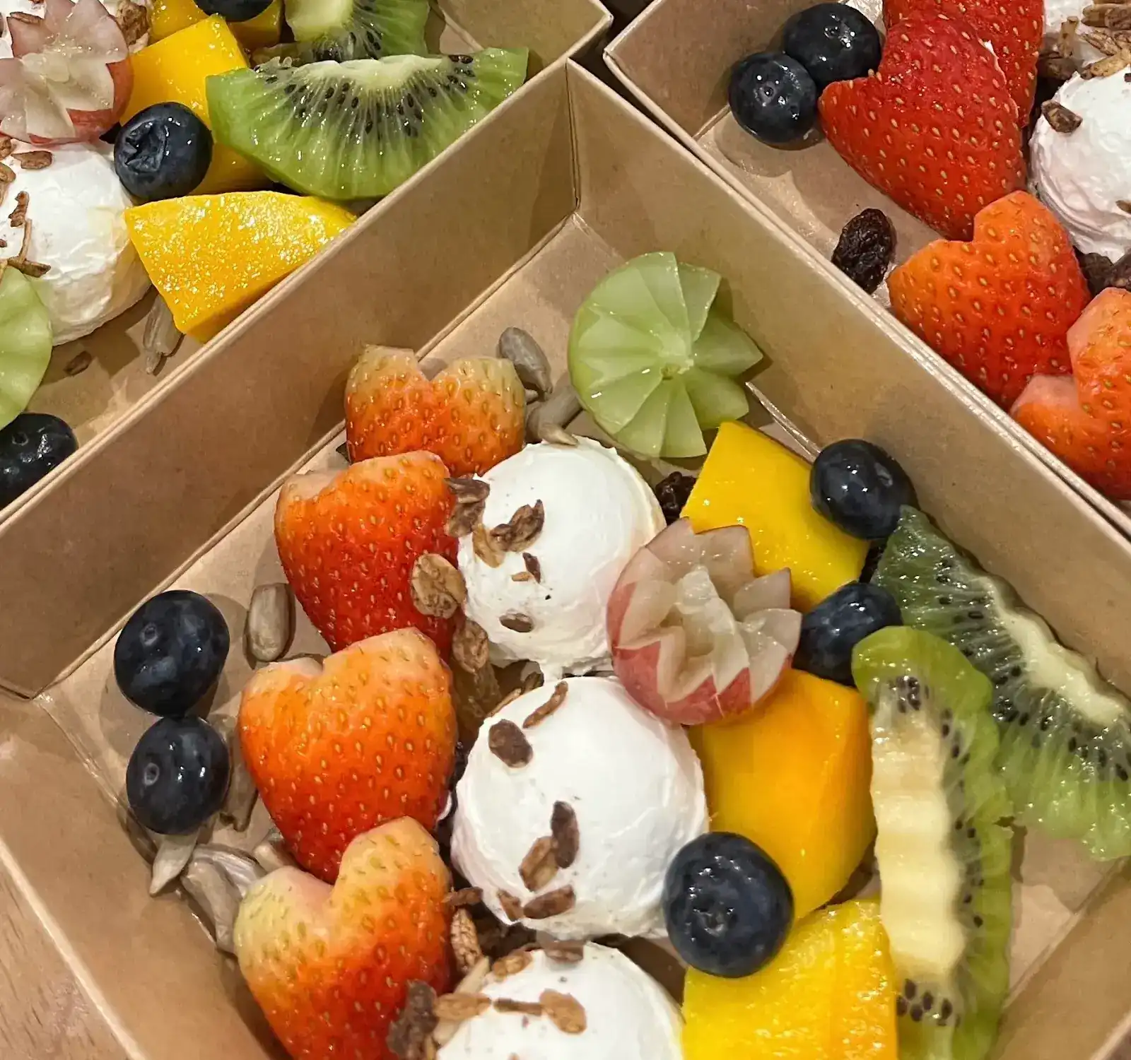 Fruit boxes with heart-cut strawberries, carved grapes, mango, kiwi, blueberries and scoops of Greek yogurt with granola
