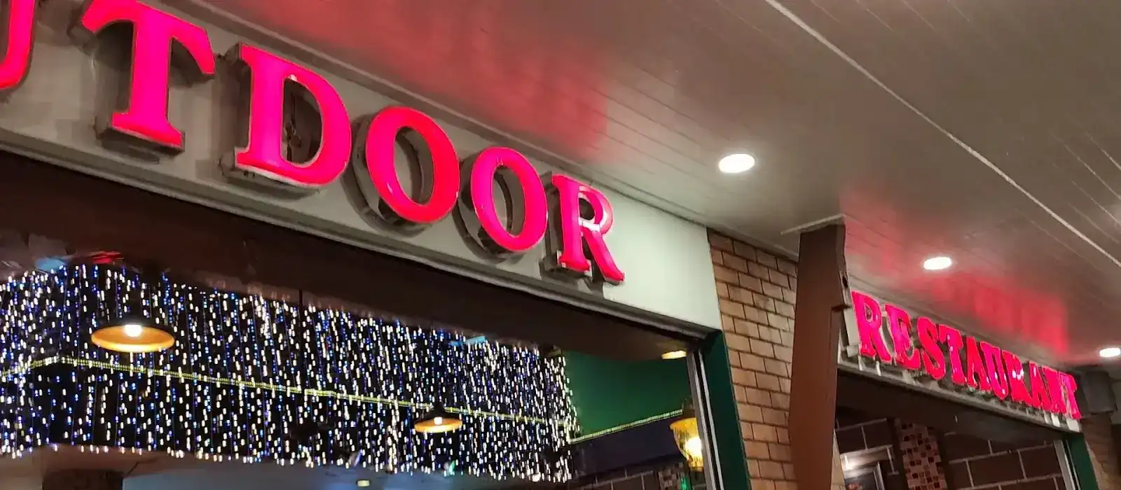 The pink OUTDOOR sign over the restaurant front at night, with fairy lights above the entrance