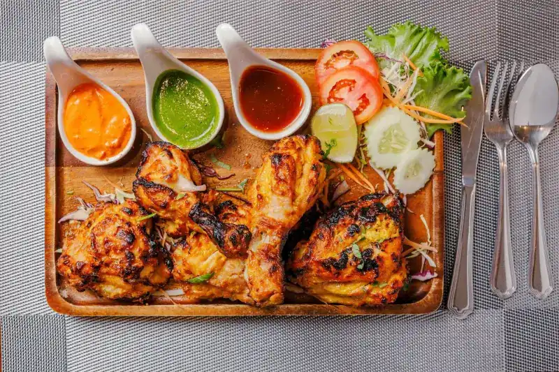 Charred tandoori chicken on a wooden board with three dipping sauces