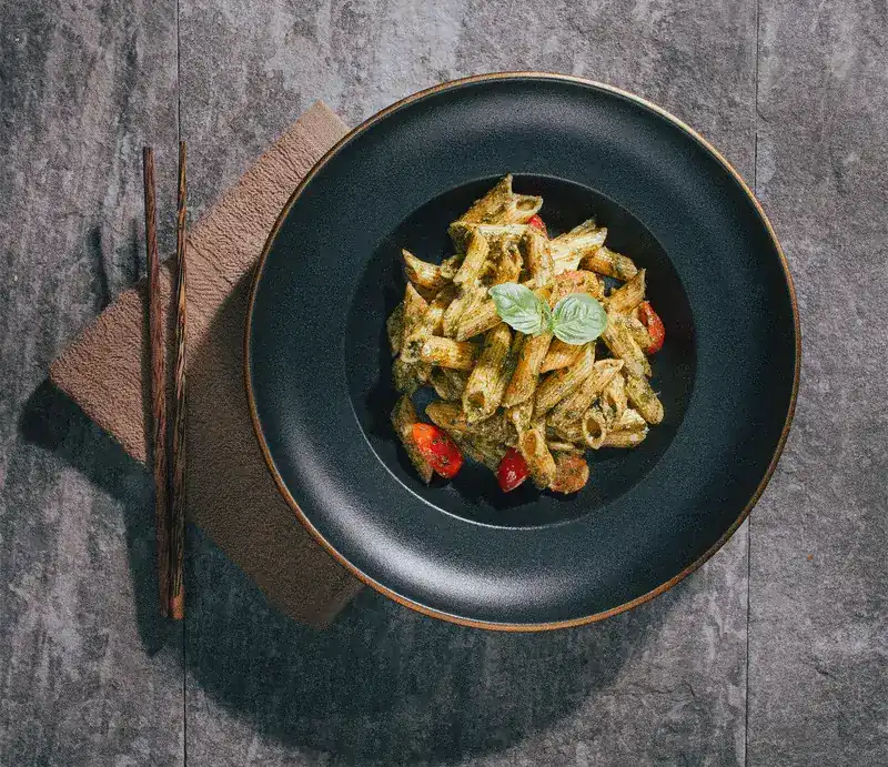 Penne pesto with cherry tomatoes and basil in a black bowl on the stone table