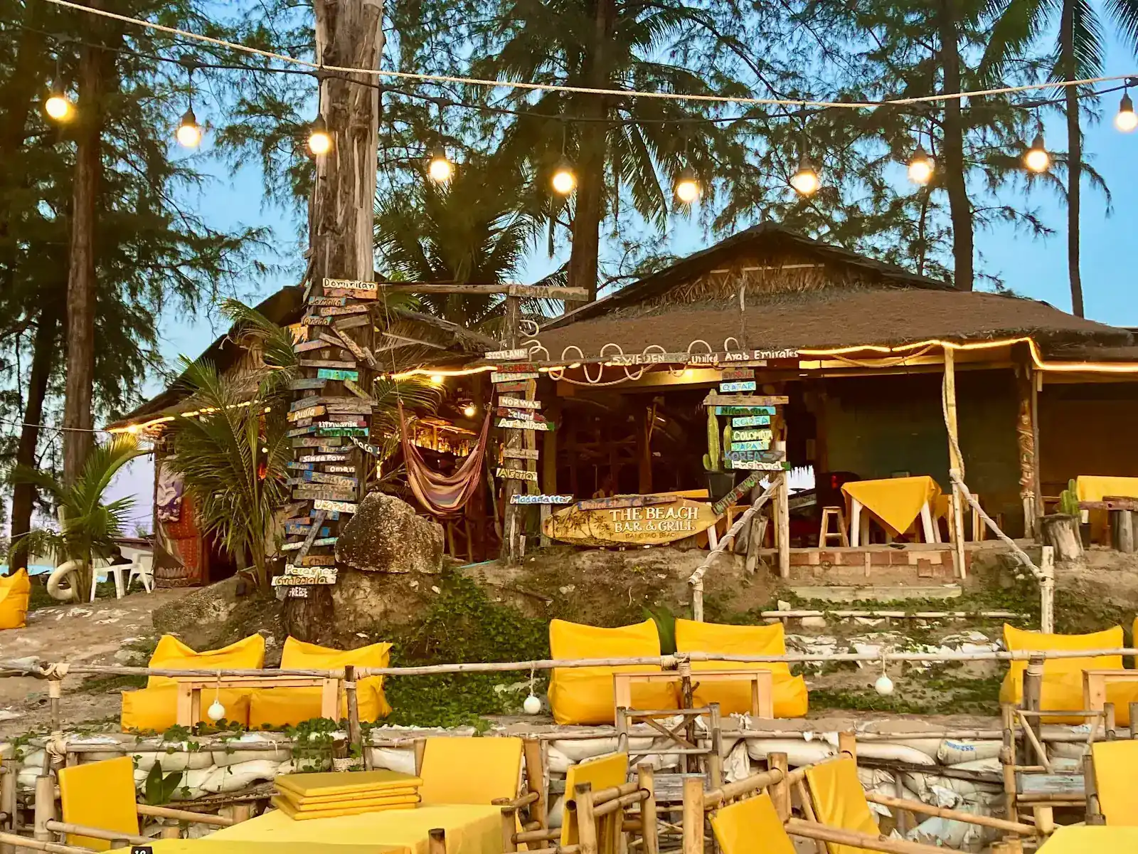 The Beach Bar & Grill at dusk: a thatched building under casuarina trees, string lights, yellow cushions on the sand and a post of hand-painted country signs.