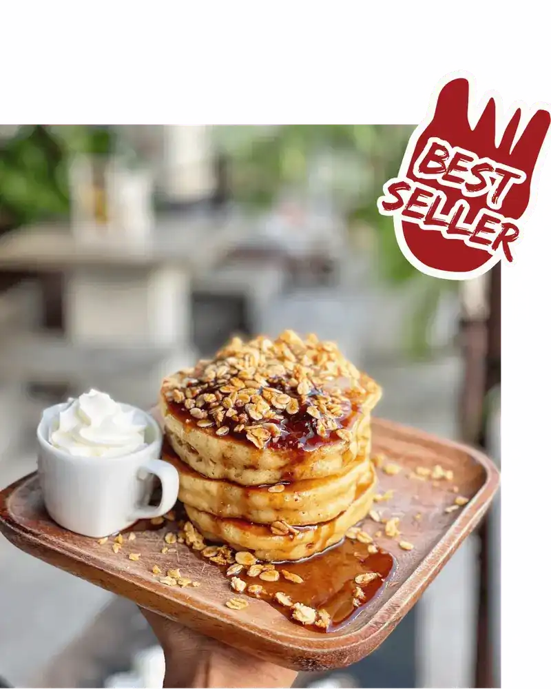 A stack of buttermilk pancakes with syrup and toasted oats, a cup of whipped cream beside it