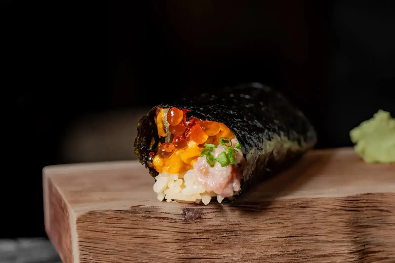 A hand roll of ikura, uni and tuna with spring onion on a wooden stand