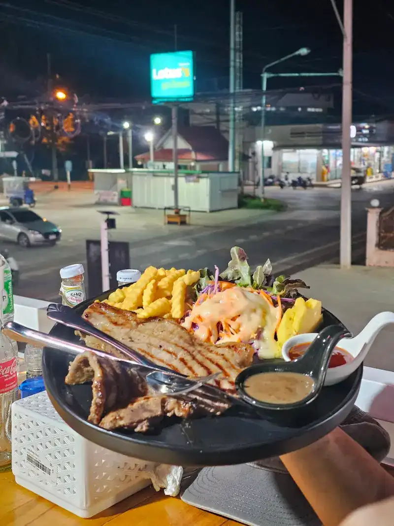A grilled steak plate with crinkle-cut fries, salad, pineapple and dipping sauces, held up over a lit road at night