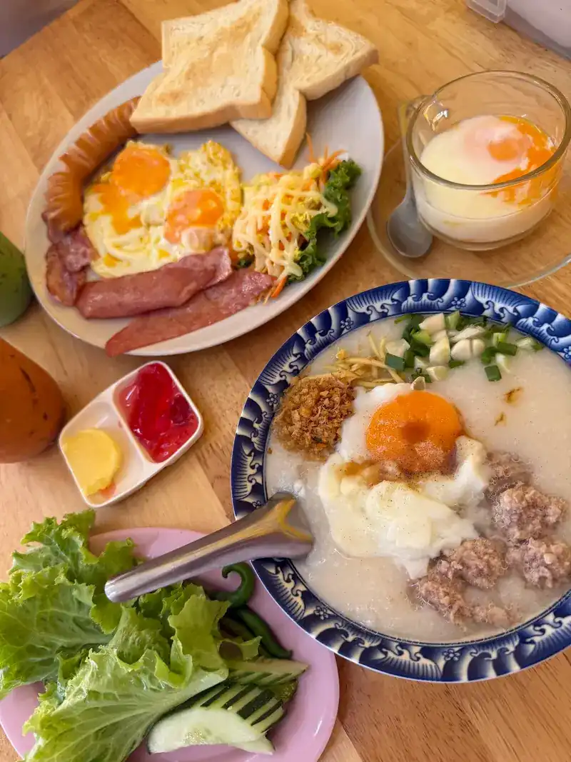 Breakfast of fried eggs, sausage, ham and toast, a bowl of congee with egg and pork, and a soft-boiled egg in a glass