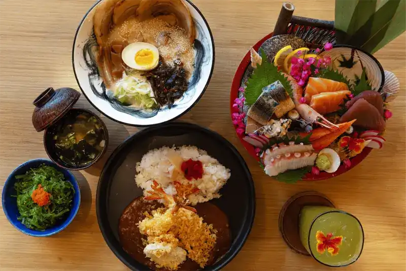 A table with a bowl of ramen, a sashimi platter, curry rice, soup and seaweed salad