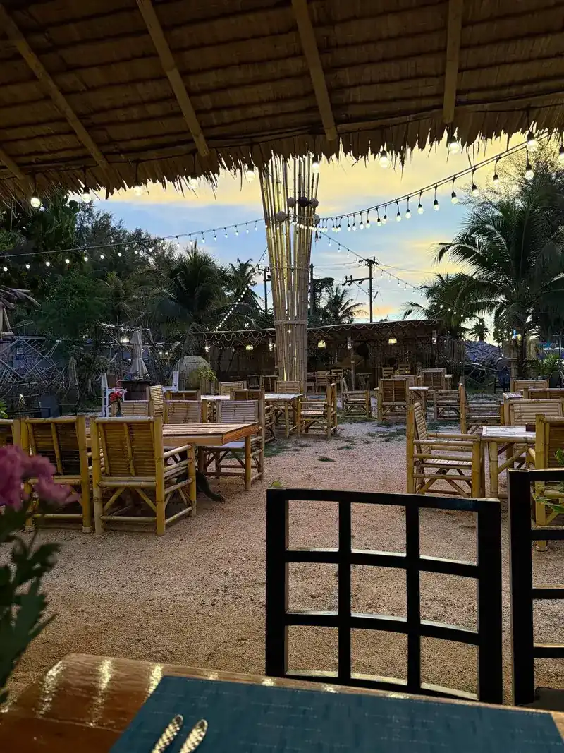 Bamboo tables and chairs on the sand, seen from under the thatched roof at sunset