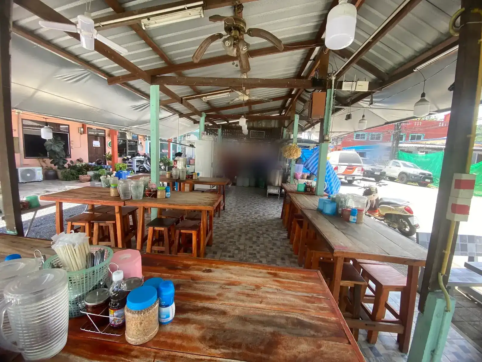 The open-sided dining room: long wooden tables and stools under ceiling fans, the noodle counter at the back