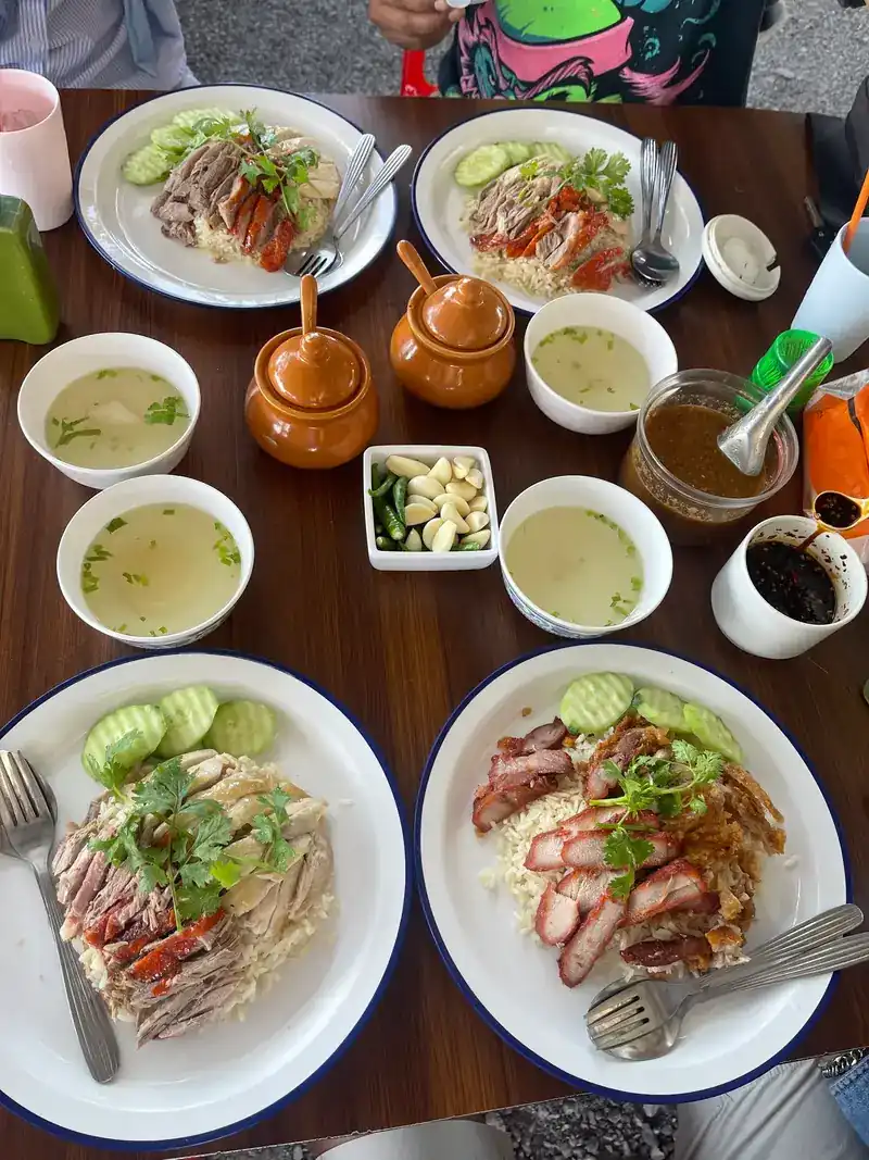 Four plates from above: chicken with roast duck, and red pork with crispy pork, with bowls of soup, garlic and chillies
