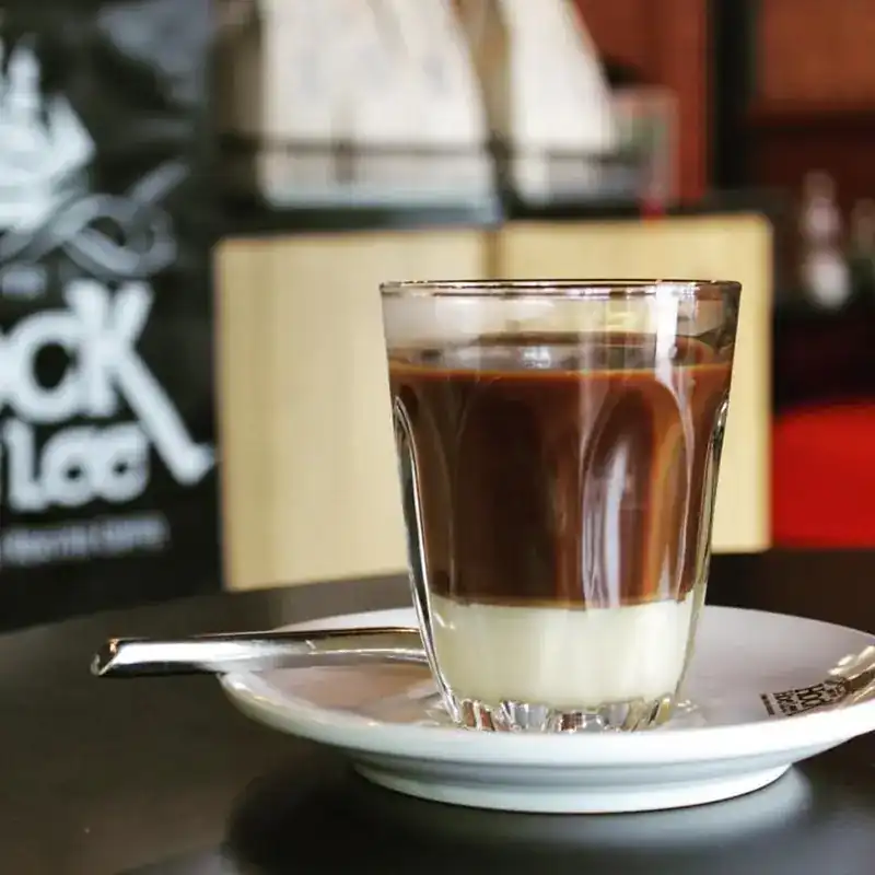A glass of black coffee layered over sweet milk on a saucer, the Hock Hoe Lee sign behind
