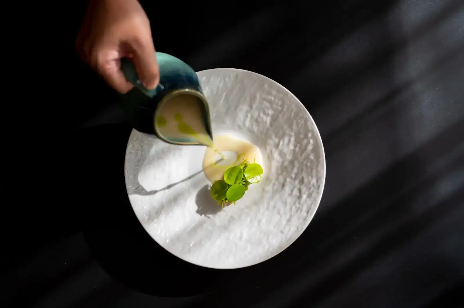 A hand pours a pale sauce from a green jug onto a white plate with small green leaves
