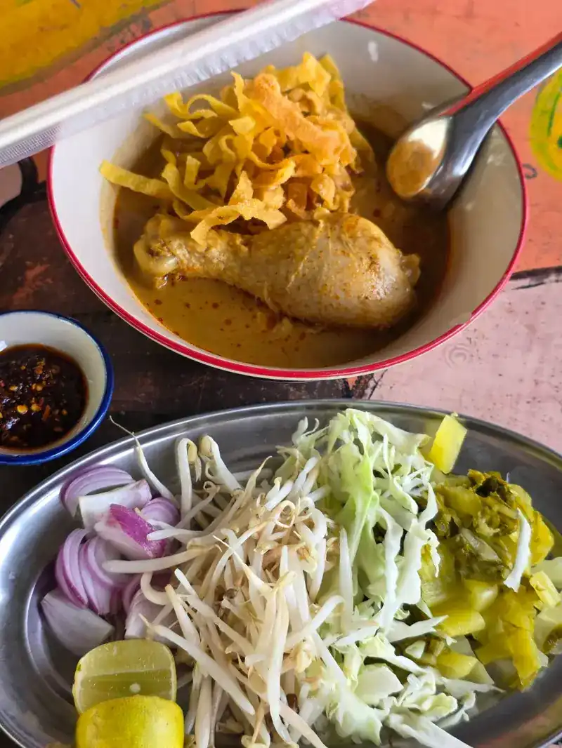 A bowl of khao soi with a chicken leg and crisp noodles, beside a plate of shallots, bean sprouts, cabbage, pickled greens and lime