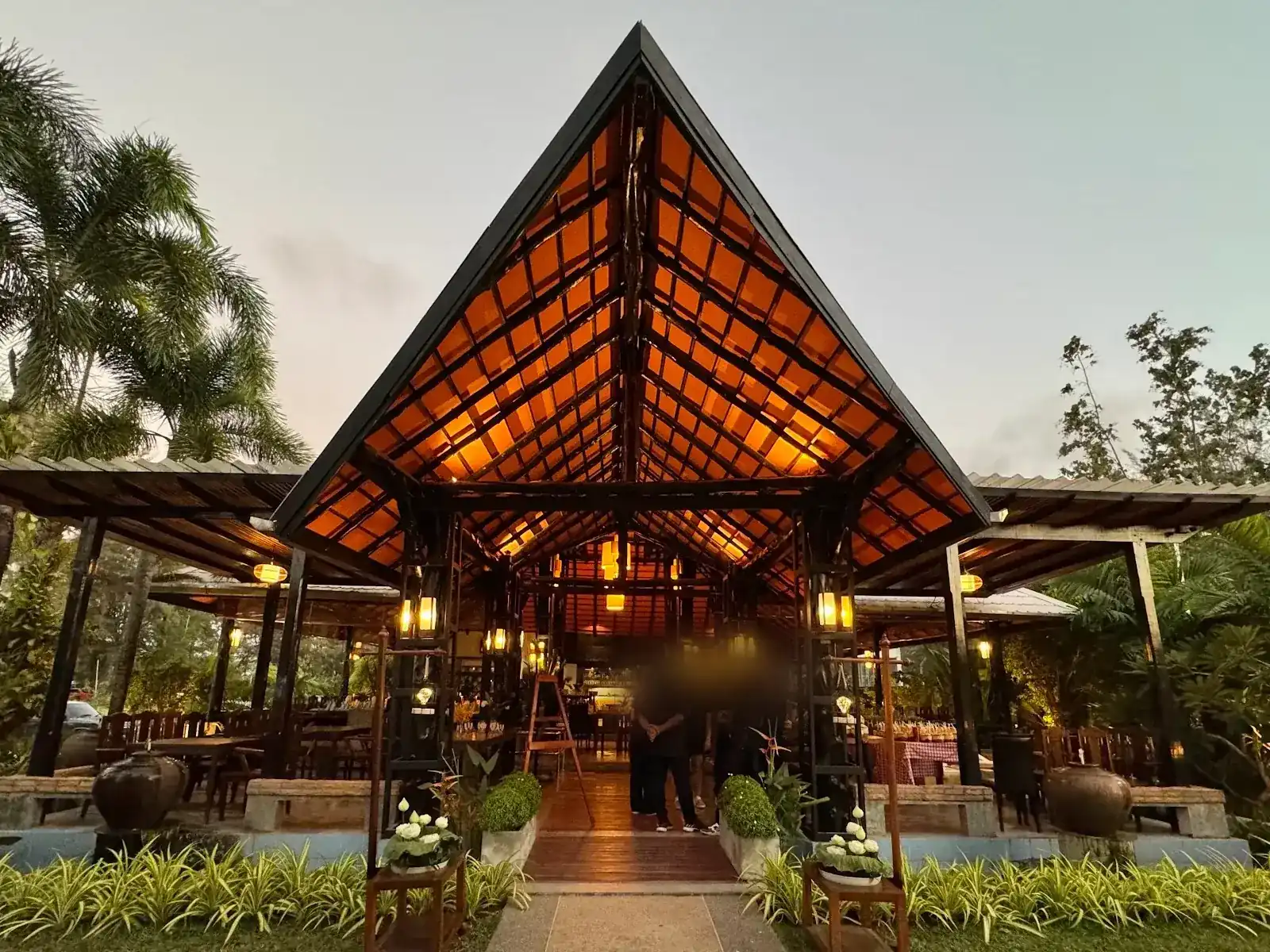 The entrance pavilion at dusk: a tall pitched roof of orange tiles on dark timber, lit from inside, with palms on either side