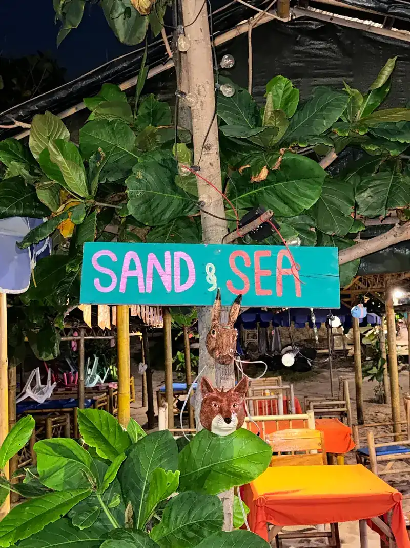 The Sand & sea sign, painted turquoise and pink, among leaves at night with tables behind it