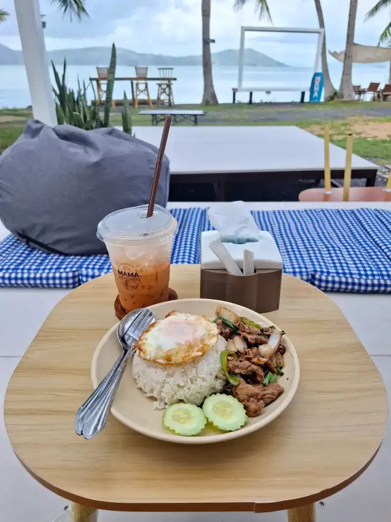 A rice plate with a stir-fry, a fried egg and cucumber, and an iced drink, at a table by the sea