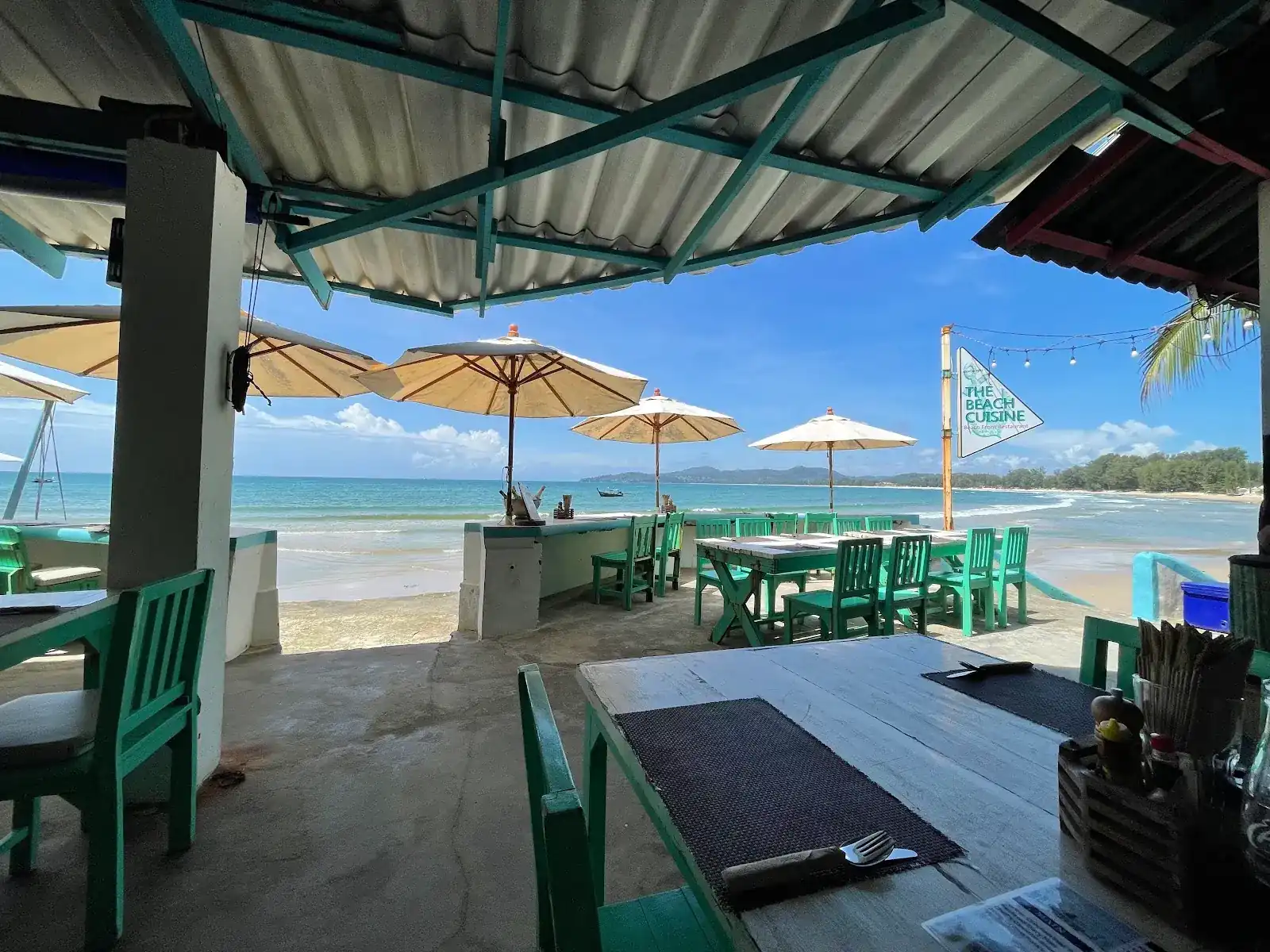 Green wooden tables under the roof, looking out over big parasols and the sea at Bang Tao Beach, with The Beach Cuisine flag on the sand