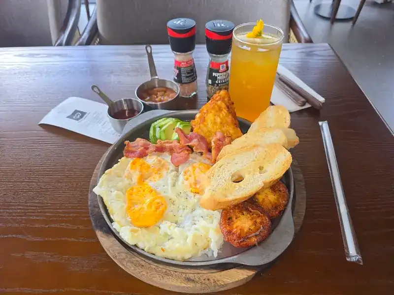 A breakfast pan of fried eggs, bacon, hash browns, toast and beans, with a glass of juice