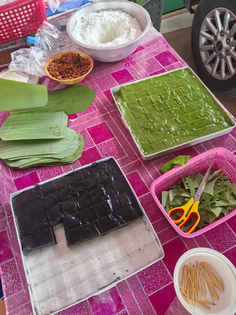 Trays of pandan-green and black khanom cut into squares, with banana leaves and scissors