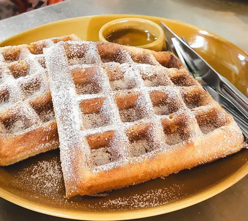 Waffles dusted with sugar, with a small cup of syrup