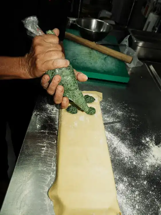 Hands piping a spinach filling onto a long sheet of fresh pasta on a floured steel bench