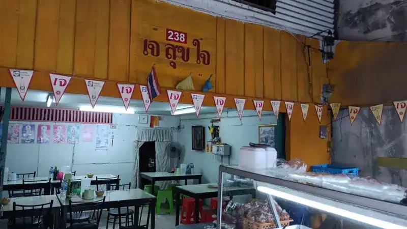 Inside the shop: tables, green and red stools, and the name เจ สุขใจ on the yellow wooden wall