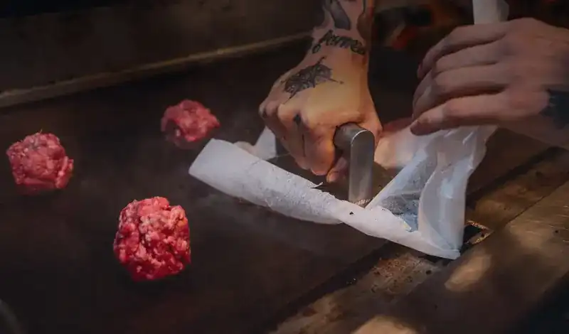 Hands pressing a ball of fresh ground beef flat on a hot griddle