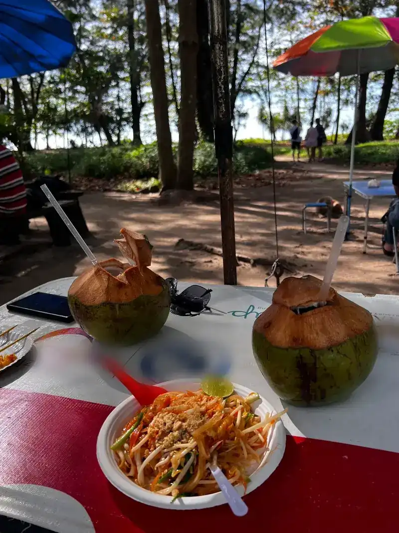 Pad thai with lime and two fresh coconuts on a table, sun umbrellas and trees behind