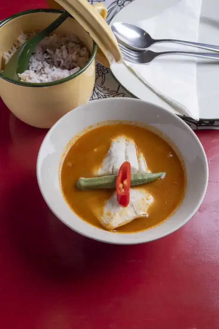 Peranakan fish curry in a white bowl on the red table, with a pot of rice