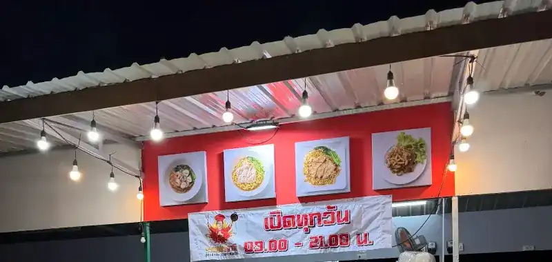 The stall at night: a red wall with four dish pictures under a tin roof, strings of bulbs, and a banner saying open every day 09.00 to 21.00 / ร้านตอนกลางคืน ผนังสีแดงมีรูปอาหารสี่รูปใต้หลังคาสังกะสี หลอดไฟเป็นแถว และป้ายเปิดทุกวัน 09.00–21.00 น.