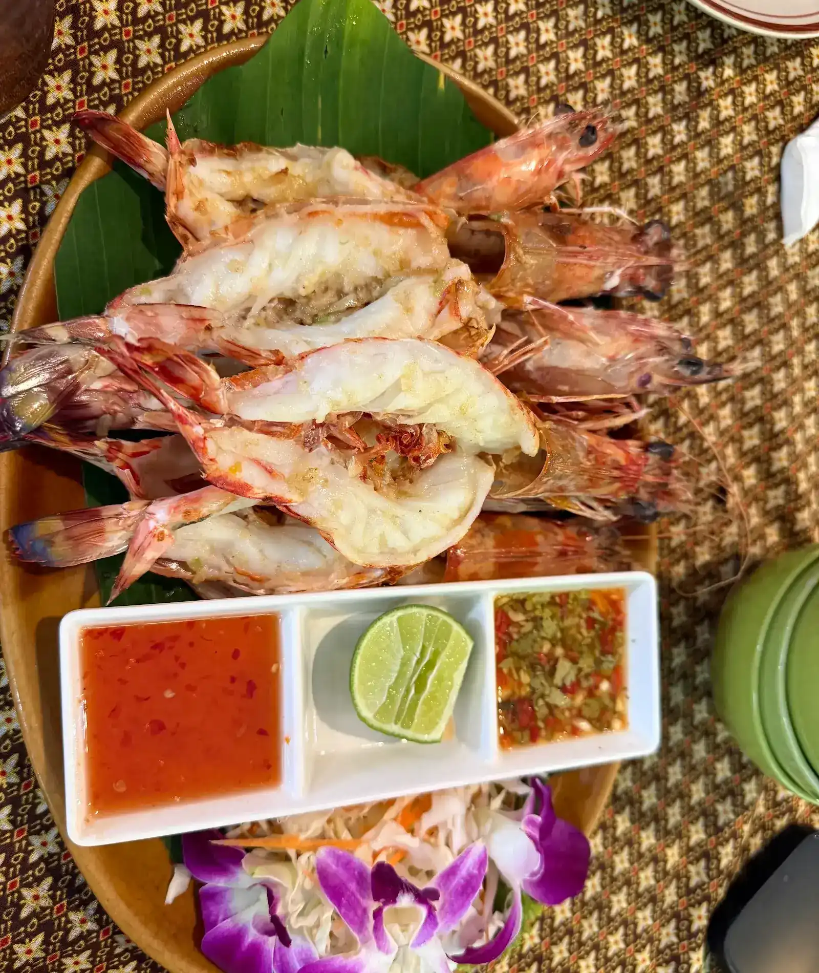 Grilled prawns, split open, on a banana leaf with two dipping sauces, lime and orchids