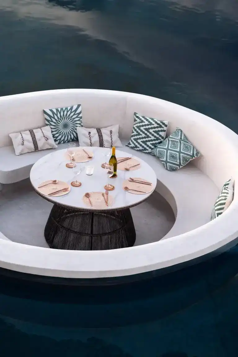 A round white sofa set like an island in dark water, its table laid for six, seen from above