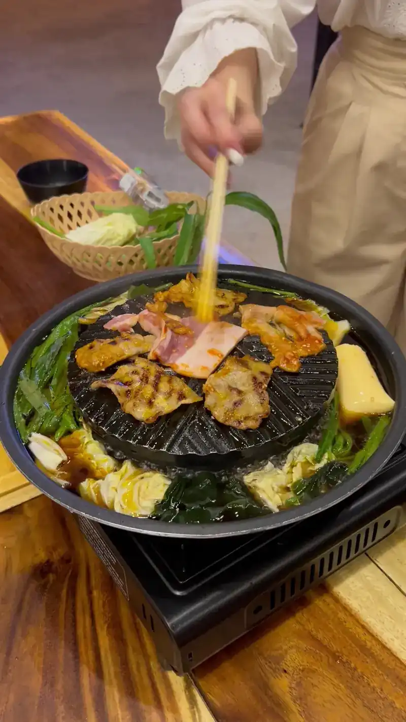 Chopsticks laying pork on the dome of a mookata pan, greens and cabbage in the broth