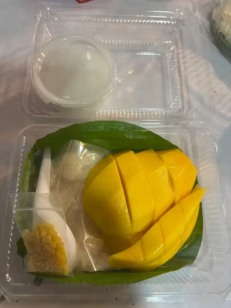 A takeaway box with a scored mango cheek and sticky rice on banana leaf, coconut cream on the side