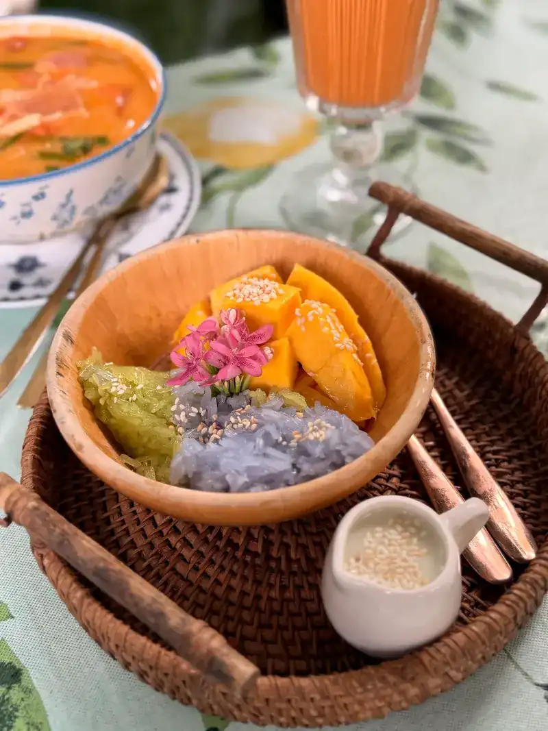 Mango with green and purple sticky rice and a jug of coconut cream on a rattan tray, a bowl of soup behind