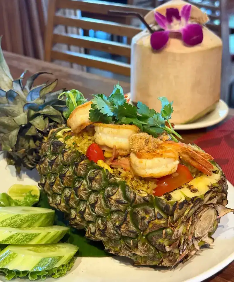 Fried rice served in a hollowed pineapple, topped with prawns, with a fresh coconut behind