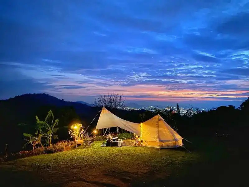 A lit tent on the camping lawn at dusk, town lights far below