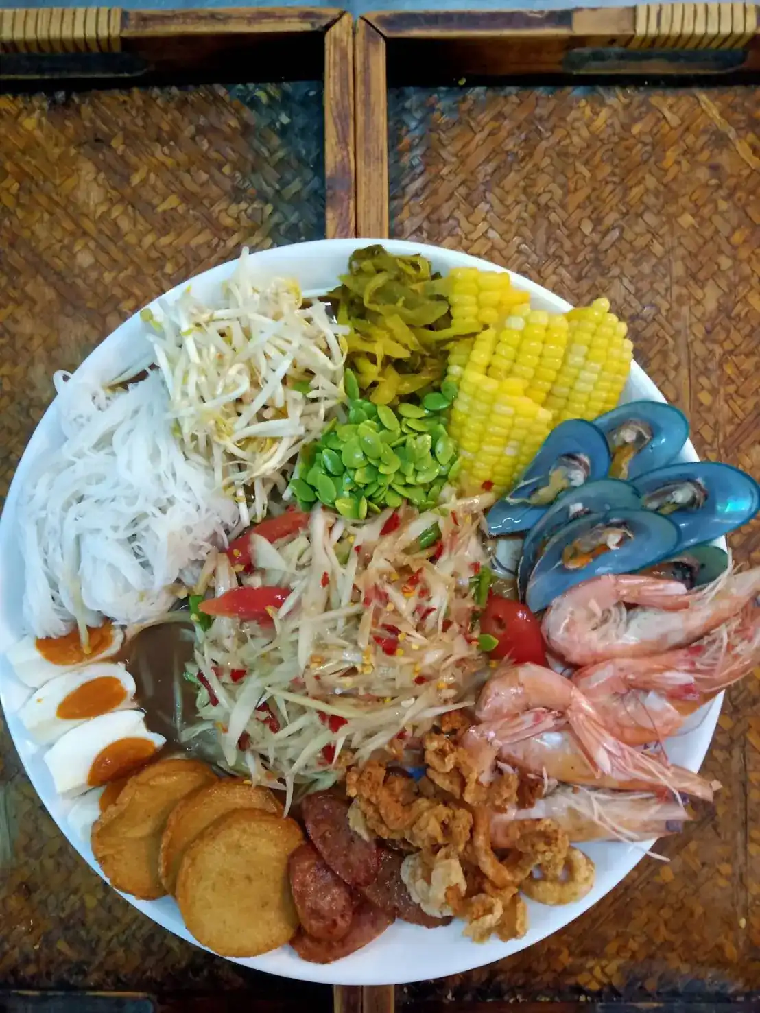 Papaya salad on a large plate with prawns, green mussels, salted egg and corn, on a woven tray