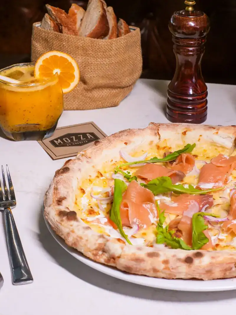 A pizza with smoked salmon, rocket and red onion beside a basket of bread