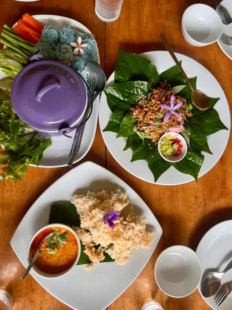 A table seen from above: a curry pot with blue rice noodles and vegetables, a dish served on fresh leaves, and a crisp fried dish with a dip
