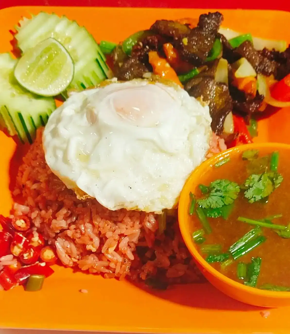 Reddish fried rice with a fried egg, a stir-fry with peppers and onion, cut cucumber, lime and a cup of soup on an orange plate