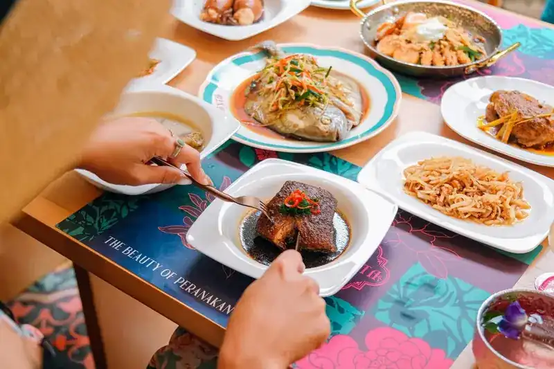 A table laid with shared dishes on a Peranakan-print placemat, hands reaching in with a fork and spoon