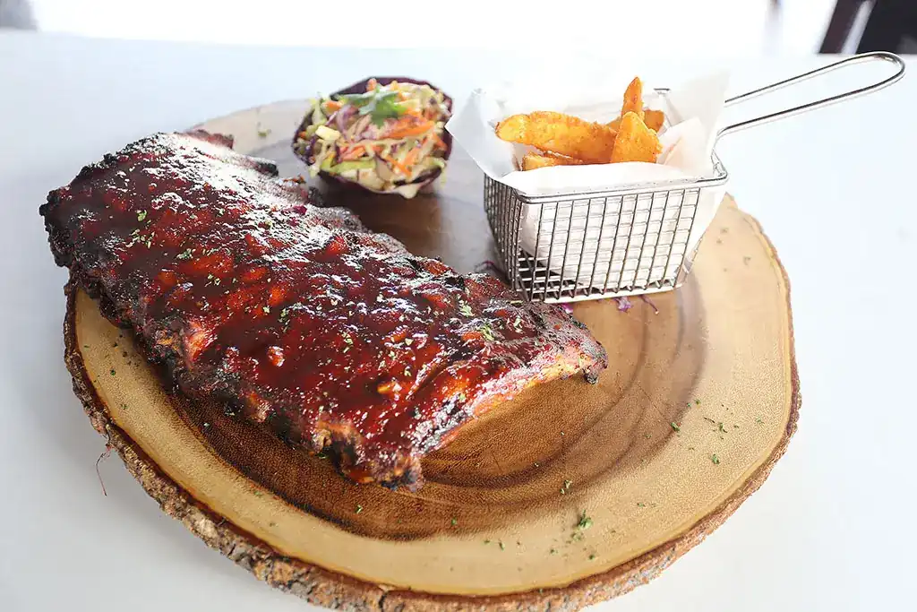 A rack of glazed BBQ pork ribs on a wooden board with coleslaw and a basket of potato fries