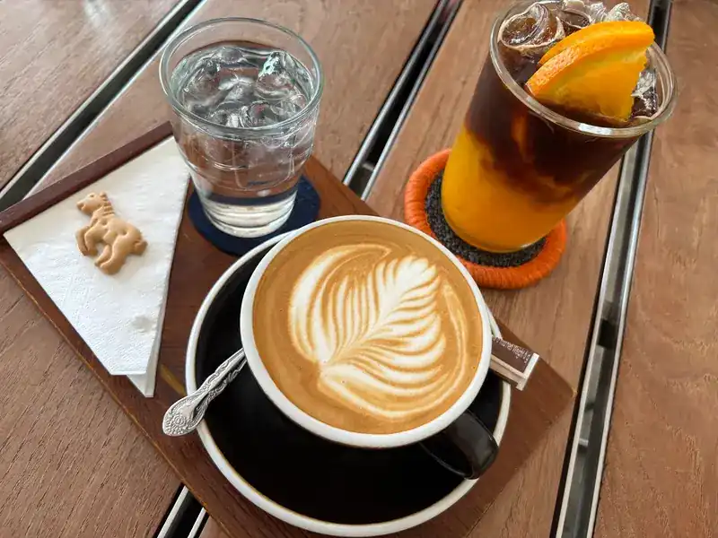 A flat white with leaf latte art beside a two-layer iced drink of black coffee over orange, on a wooden table