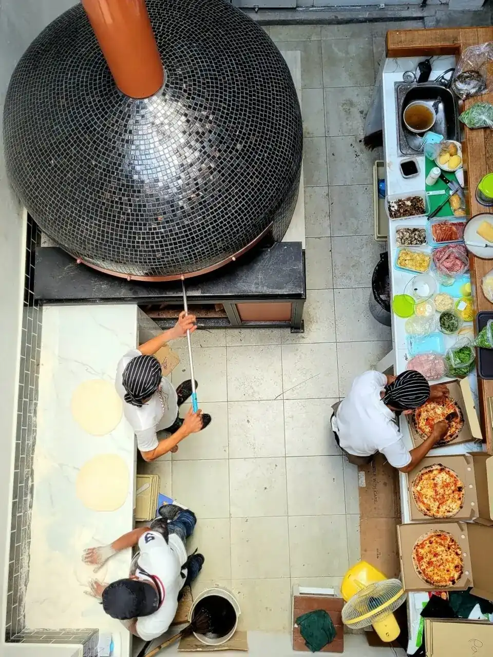 A black mosaic wood oven with a copper flue, seen from above, with pizza makers at work