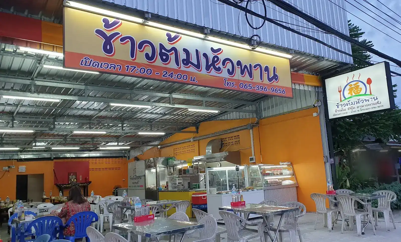 The front of Huaphan Rice Porridge Shop at dusk: a yellow sign with the shop name, opening hours and phone above steel tables, orange walls and the kitchen counter under a tin roof